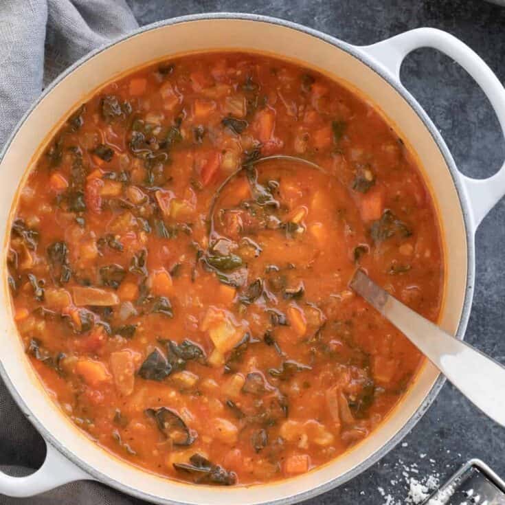 Hearty vegetable soup with tomatoes, carrots, and greens in a white pot on a dark surface.