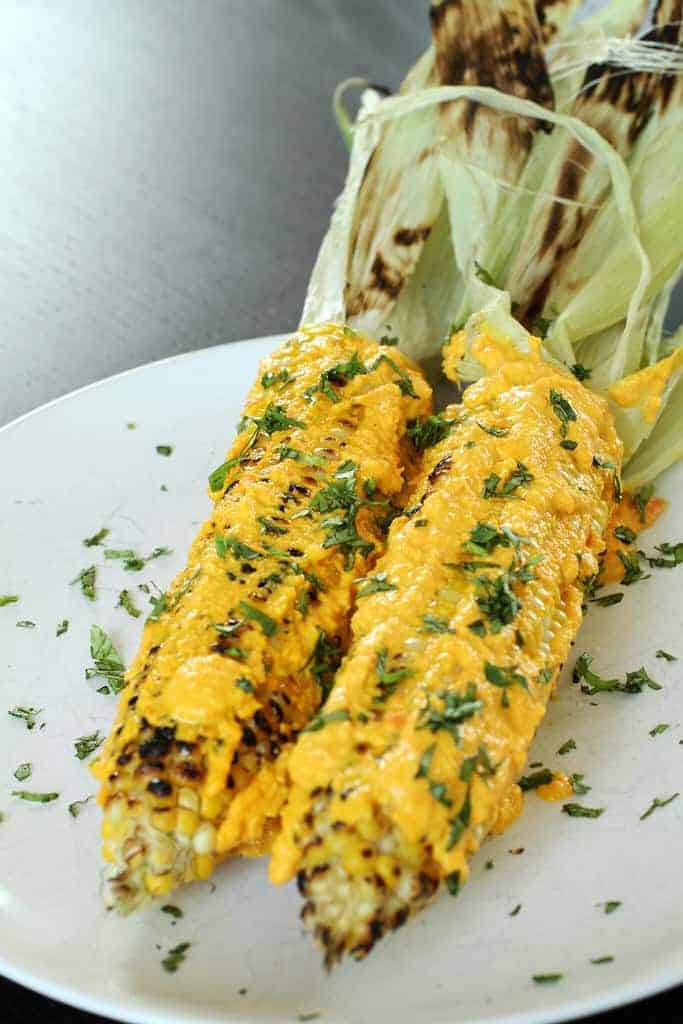 Grilled corn on the cob with cheesy topping and fresh herbs on white plate.