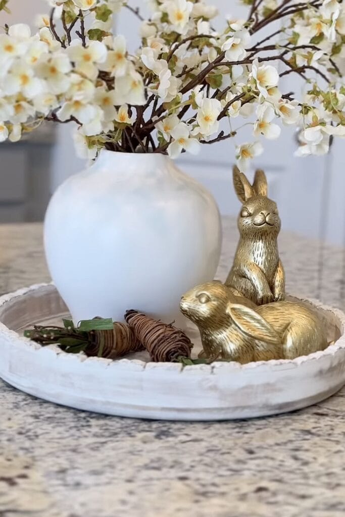 A white vase filled with blooming white artificial flowers on a round tray with gold bunny and rabbit figurines for Easter or spring decor.