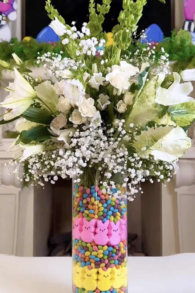 A beautiful bouquet of white roses, lilies, and baby's breath in a glass vase filled with colorful candy-coated bunny and chick decorations for spring or Easter celebrations.