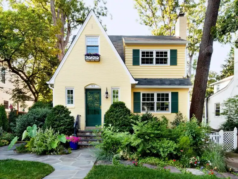 Yellow painted brick house with green shutters and lush garden.