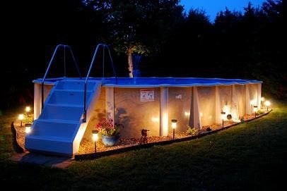 Illuminated above-ground swimming pool at night with surrounding garden and landscape.