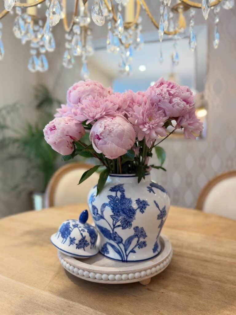 Pink peonies in a blue and white vase on a wooden table with chandelier overhead.