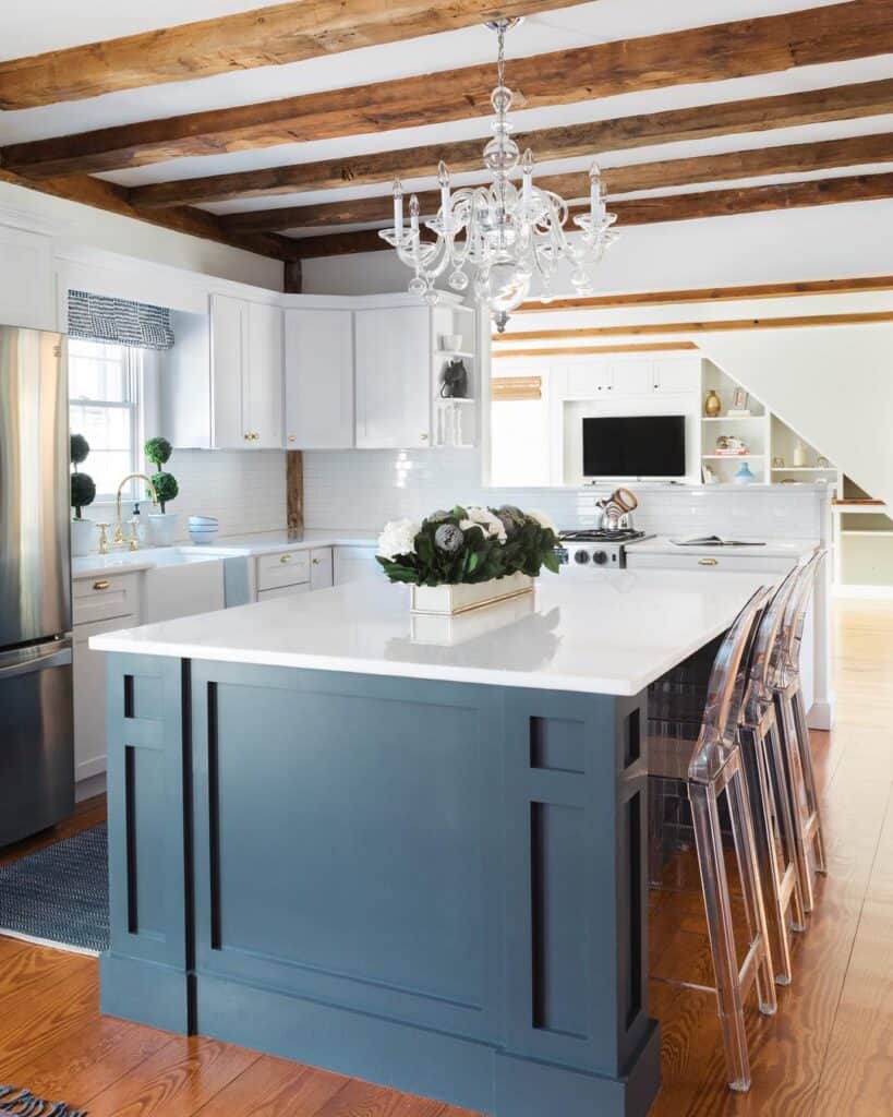Bright kitchen with blue island, white cabinets, chandelier, and wooden ceiling beams.