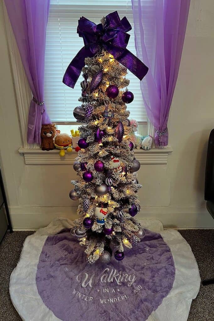 Decorated purple Christmas tree with ornaments and bow in a cozy room.