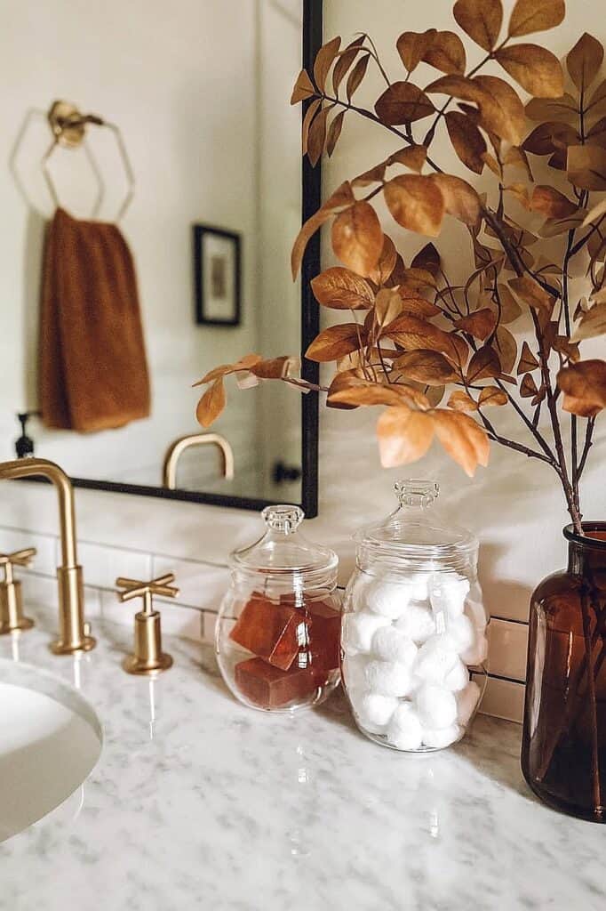 Autumn-inspired bathroom decor with dried leaves and glass jars on marble countertop.