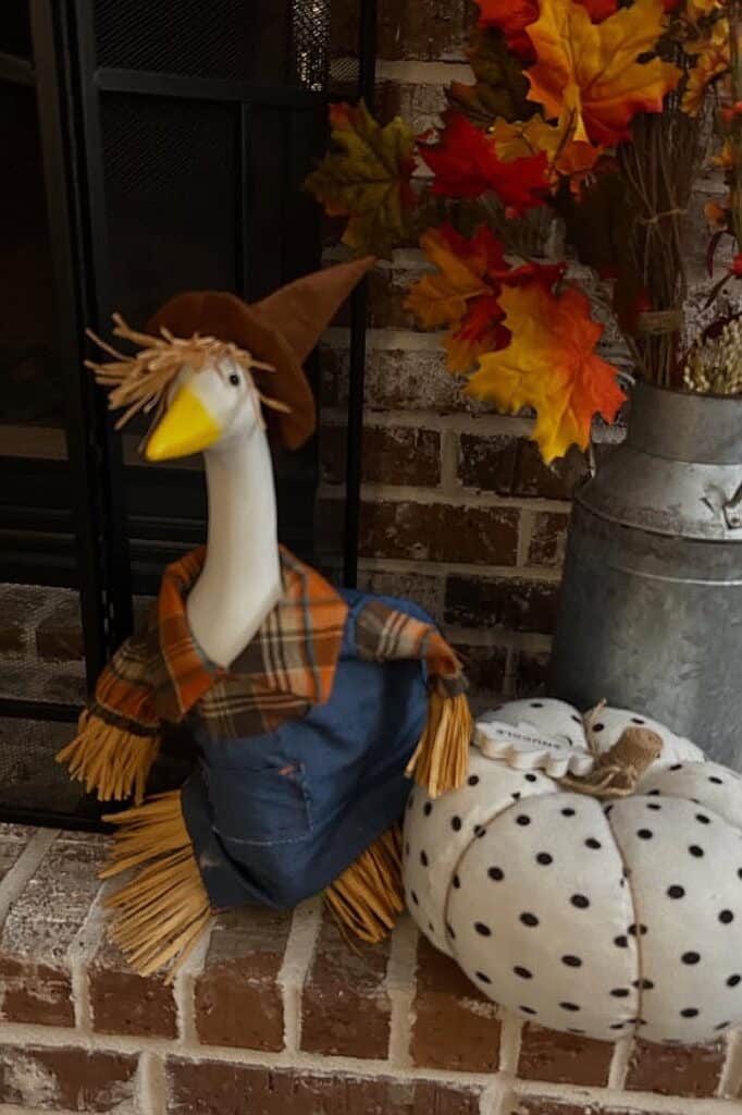Decorative fall porch scene with a goose figurine, pumpkins, and autumn leaves.