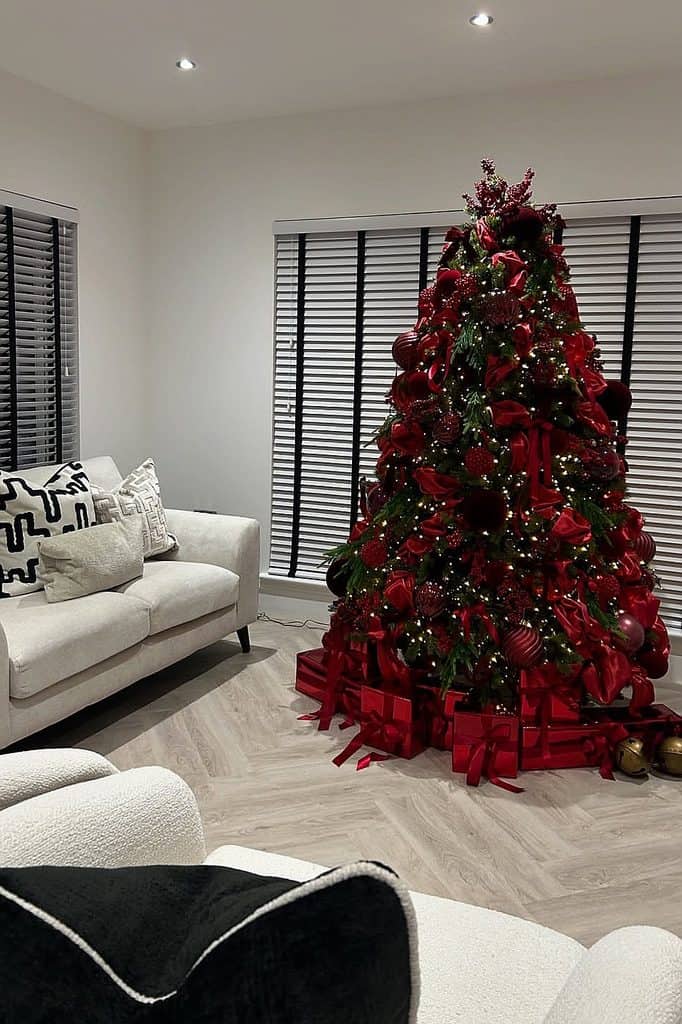 Beautifully decorated red Christmas tree with ornaments and gifts in a modern living room.