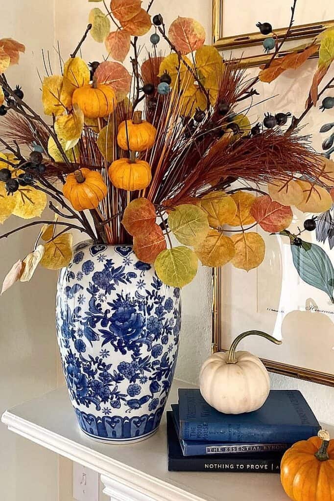 Fall floral arrangement with pumpkins and dried leaves in a blue and white vase.