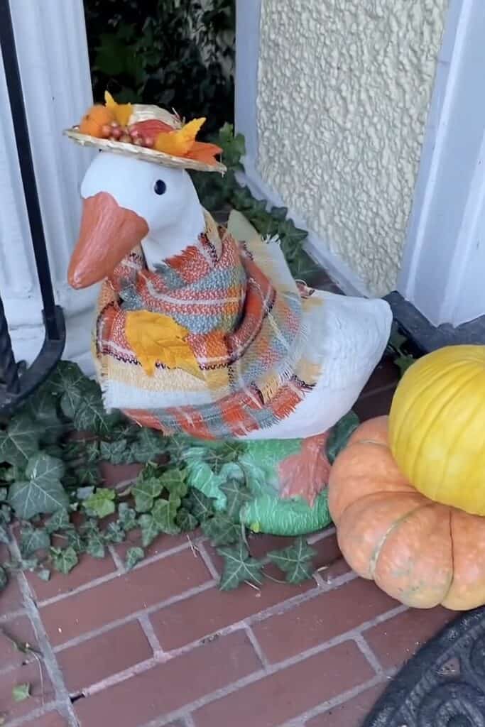 Decorative goose with fall leaves and pumpkins on porch for autumn decor.