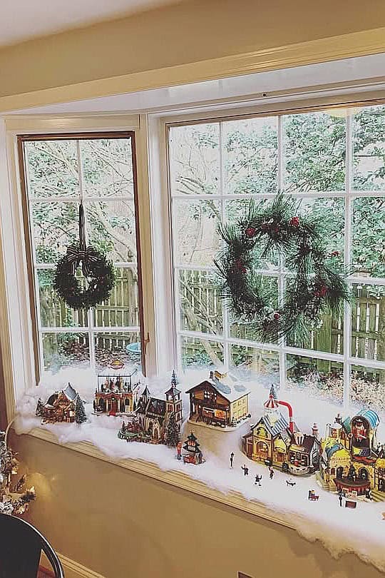 Festive Christmas village scene with wreaths and holiday decorations on windowsill.