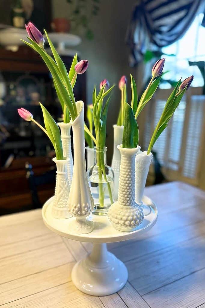 Milk glass vases with pink tulips on a white cake stand in a cozy home setting.