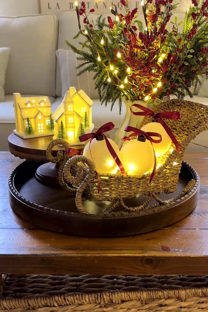 Festive Christmas village display with illuminated houses, flowers, and a wicker sleigh.