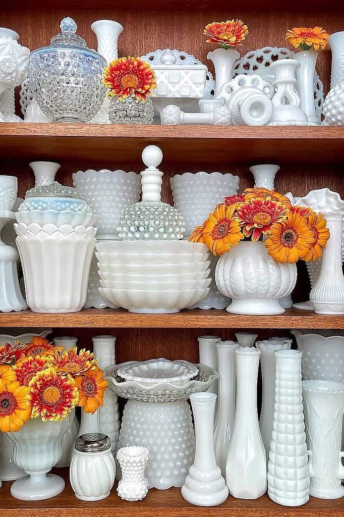 Display of white milk glass vases and bowls with orange flowers on wooden shelves.