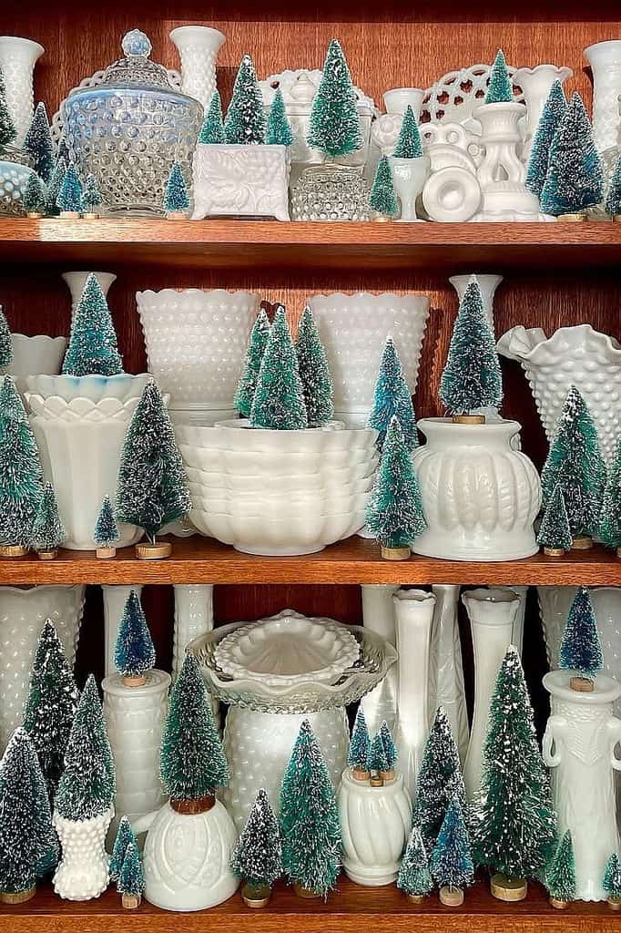 Assorted milk glass vases and bowls with miniature Christmas trees for holiday decor.