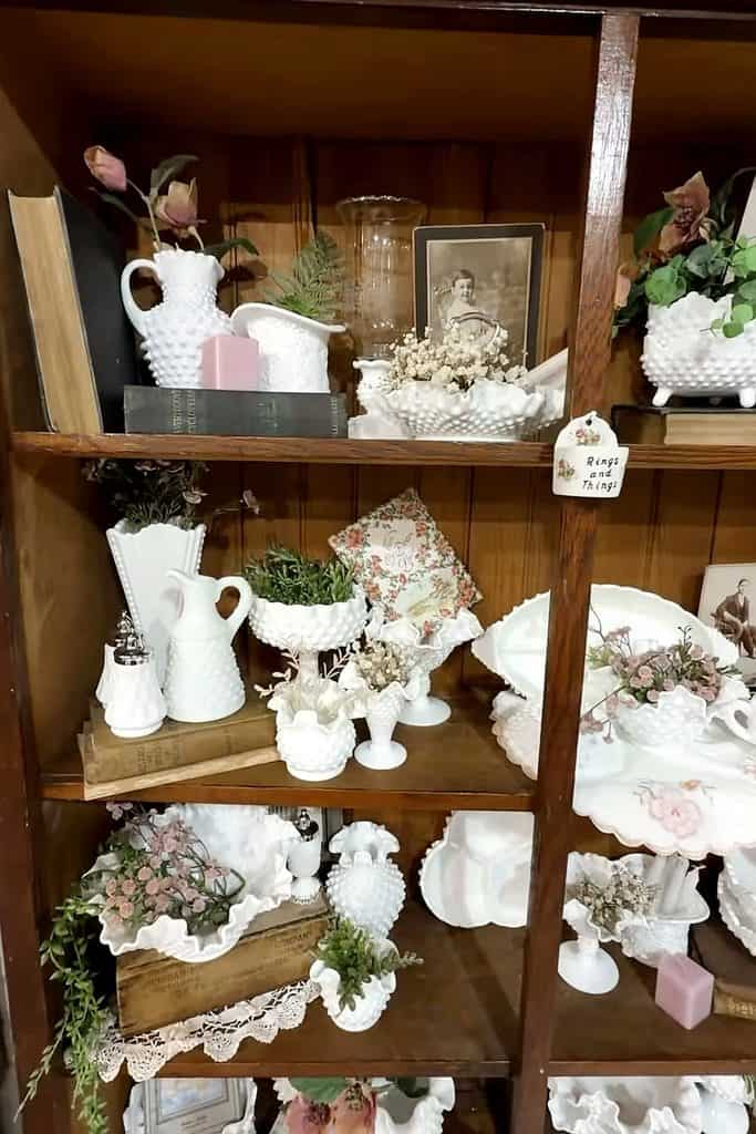 Vintage milk glass collection with flowers and decorative items on wooden shelves.