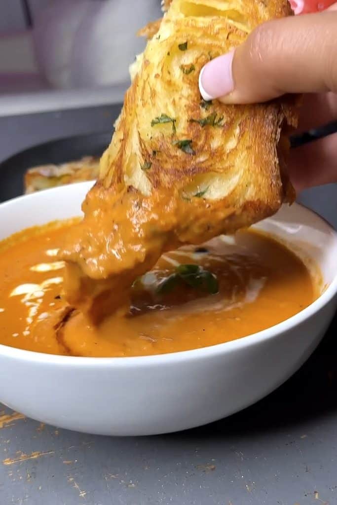 Hand dipping cheesy bread into warm tomato soup for a cozy meal.