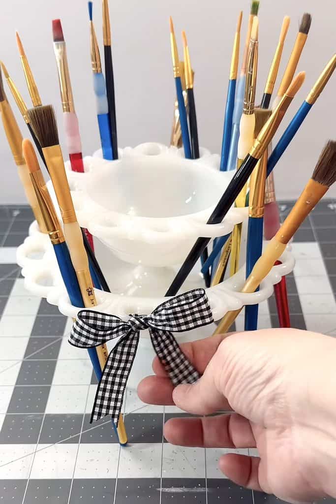 Colorful paintbrushes in a white ceramic holder with a black and white checkered ribbon.